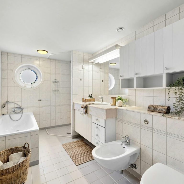 Baño moderno con bañera, lavabo y bidet, decorado con azulejos claros y plantas.